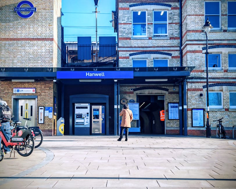 A person standing outside the entrance of Hanwell station, part of the Elizabeth Line, in a brick building with large windows and blue signage displaying 'Hanwell'. The scene features a paved walkway