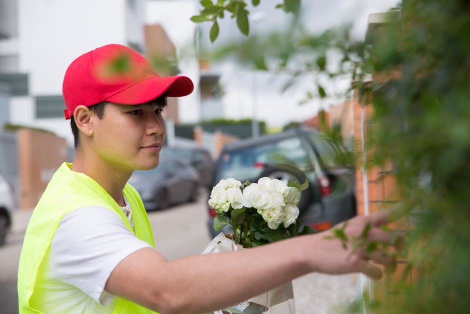 A delivery person wearing a red cap and a high-visibility yellow vest is placing a bouquet of fresh white hydrangeas wrapped in clear plastic beside a brick wall outdoors. The hydrangeas have lush gre